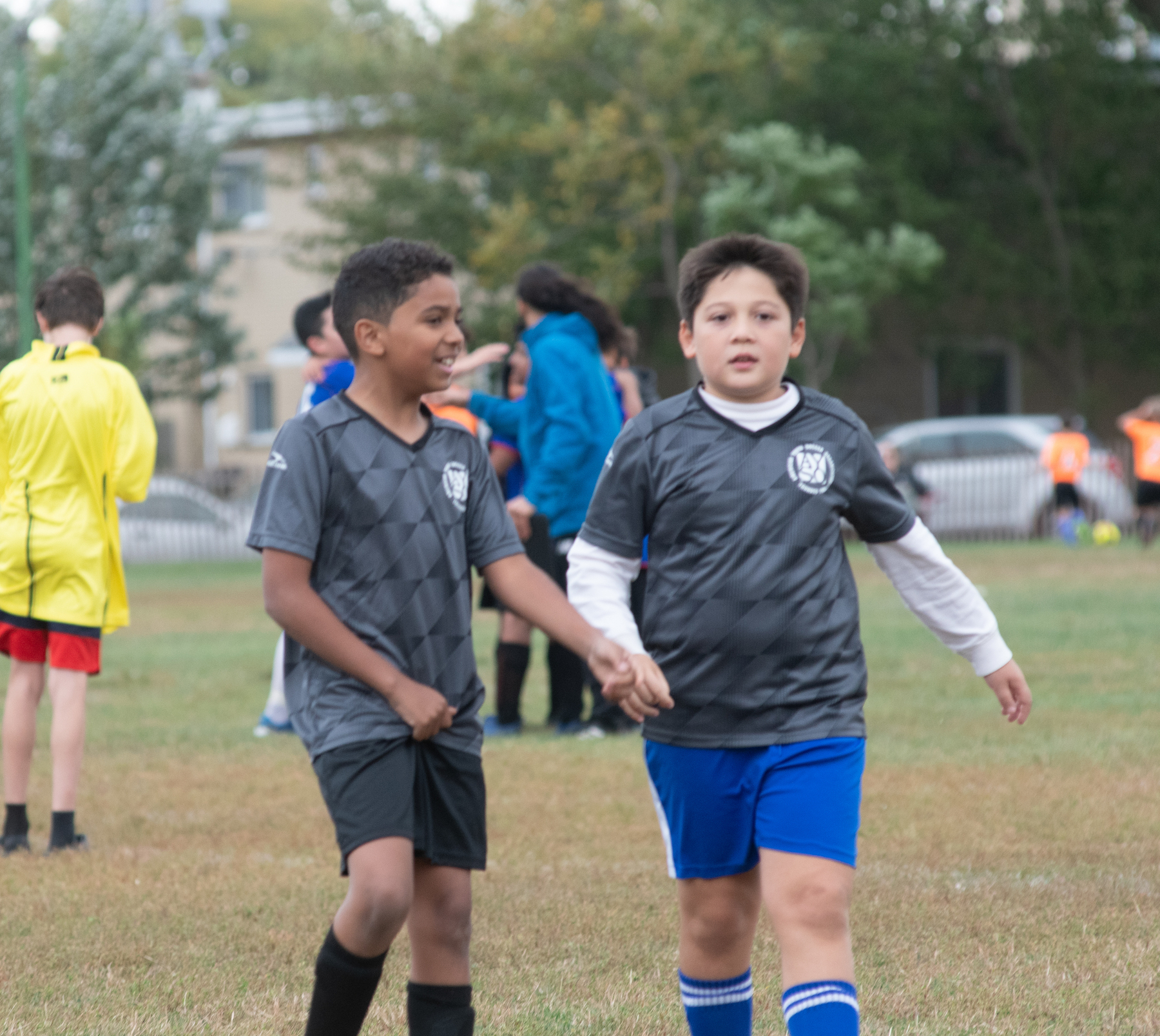 Two young boys in soccer uniforms walk on a grassy field with others in the background.